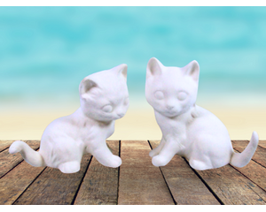 2 small ready to paint cat figurines sitting on a rustic wooden table by the ocean.  The cats are facing each other, head facing the camera.